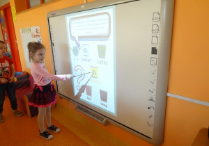 Dziewczynka stoi pod tablicą interaktywną i wskaźnikiem pokazuje kosz na odpady plastikowe.